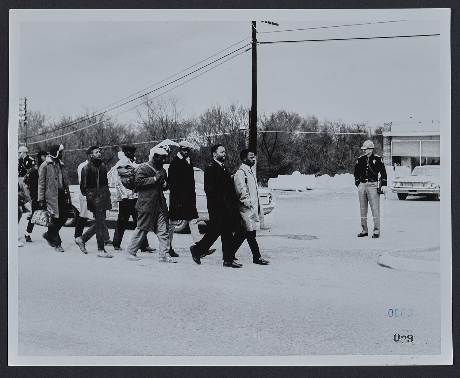 Black and white photograph of John Lewis in a tan trench coat and backpack leading civil rights marchers