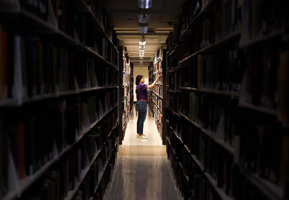 A Library staff member selects a book in the background while the lights stacks in the foreground remain dimmed.